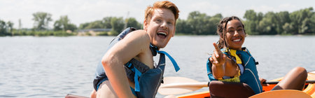 young and excited redhead man with open mouth looking at camera near overjoyed african american woman in life vest pointing with finger while sailing in kayak on summer day, bannerの写真素材