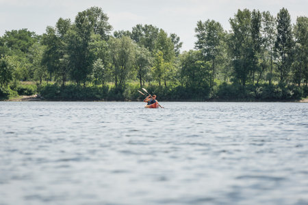 view from afar of active and interracial couple in life vests sailing in sportive kayak during recreation weekend on river with green bank during water recreation in summerの写真素材
