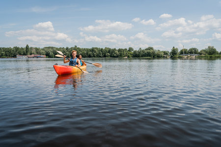 active multiethnic couple in safe vests sailing in sportive kayak on picturesque lake with green shore under blue and cloudy sky during water recreation on summer weekendの写真素材