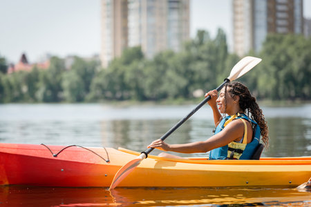 side view of active and charming african american woman in life vest sailing in sportive kayak with paddle on summer weekend day on blurred backgroundの写真素材