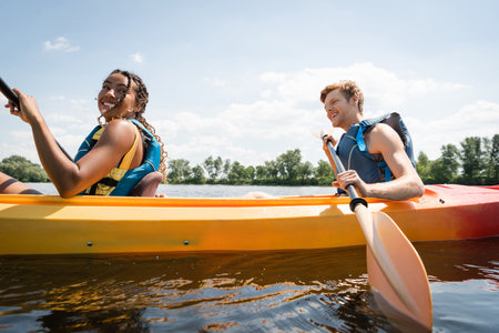 smiling and pretty african american woman sailing in sportive kayak with active redhead man in life...