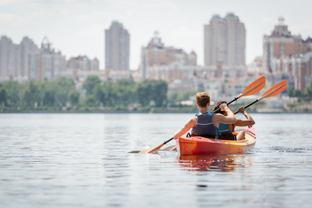 back view of active multiethnic couple in life vests paddling in sportive kayak on calm water surface on summer day with view of blurred urban buildings on riversideの写真素材