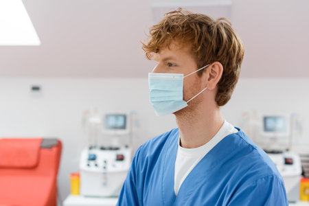 profile of redhead, young and positive doctor in medical mask and blue uniform looking away in blood...