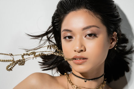 top view of young asian woman with short brunette hair holding golden jewelry in mouth while looking at camera and lying on grey background, everyday makeup, wet hairstyleの写真素材