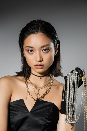 portrait of brunette and asian young woman with short hair holding golden and silver jewelry while wearing glove and standing in strapless dress on grey background, wet hairstyle, natural makeupの写真素材