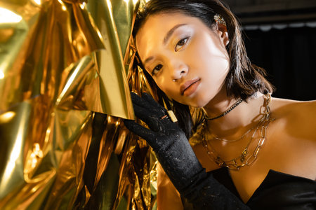 portrait of alluring asian young woman with wet short hair and black glove touching shiny yellow background, model, looking at camera, wrinkled golden foil, natural asian beautyの写真素材