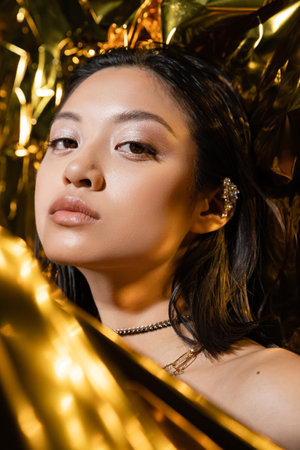 portrait of beautiful asian young woman with wet short hair posing next to shiny golden background, model, looking at camera, wrinkled yellow foil, natural asian beautyの写真素材