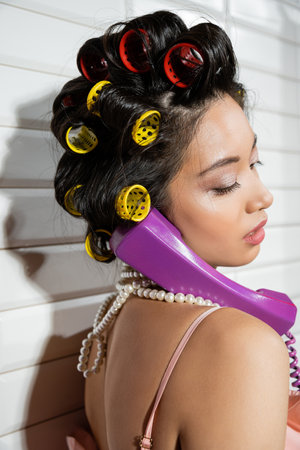 fashionable and asian young woman with hair curlers and pearl necklace talking on purple retro phone near white tiles, housewife, retro fashion, vintage-inspiredの写真素材
