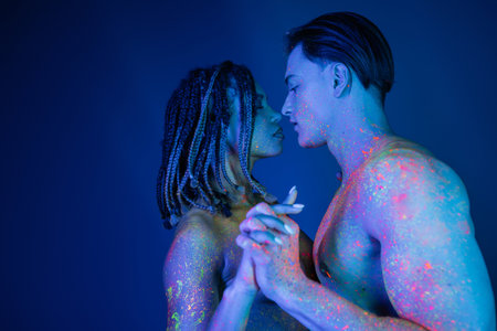 side view of interracial couple in colorful neon body paint standing with clenched hands, african american woman with dreadlocks and shirtless muscular man on blue background with cyan lightingの写真素材