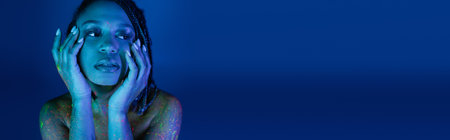 portrait of youthful african american woman with dreadlocks, in colorful neon body paint, holding hands near face and looking away on blue background with cyan lighting effect, bannerの写真素材