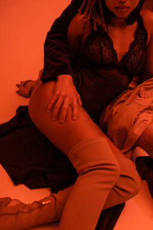 partial view of young man embracing leg of sexy african american woman in over knee boots and black lace bodysuit sitting on clothes on orange background with red lighting effectの写真素材