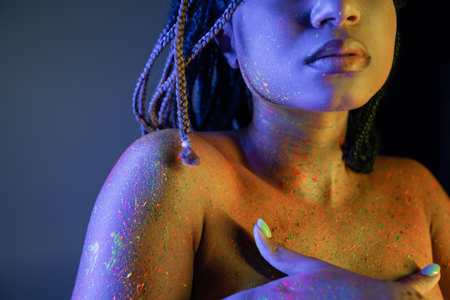 partial view of young and bare-chested african american woman in radiant and colorful neon body paint covering breast with hands on blue background with yellow lighting effectの写真素材
