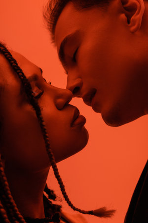 close up view of young and sexy interracial couple in love, young man and african american woman with dreadlocks kissing with closed eyes on orange background with red lighting effectの写真素材