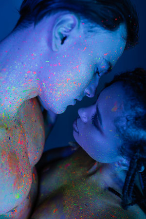 intimate moment of young multicultural couple in colorful neon body paint, bare-chested man and sexy african american woman on blue background with cyan lightingの写真素材