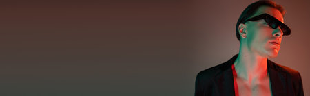 portrait of glamorous bare-chested man with brunette hair posing in black blazer and dark trendy sunglasses on grey background with red lighting and copy space, bannerの写真素材