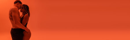 side view of young shirtless man in black pants embracing provocative african american woman in lace bodysuit on orange background with red lighting effect, bannerの写真素材