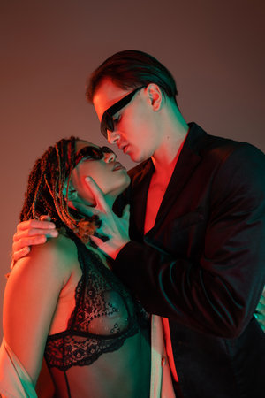 young and self-assured man in dark sunglasses and black blazer seducing enchanting african american...