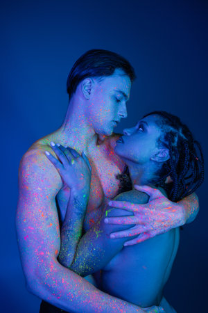 youthful interracial couple in colorful neon body paint embracing on blue background with cyan lighting, shirtless man with muscular torso and african american woman with dreadlocksの写真素材