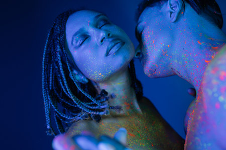 sexy interracial couple in colorful neon body paint, african american woman with dreadlocks and closed eyes near young bare-chested man on blue background with cyan lightingの写真素材