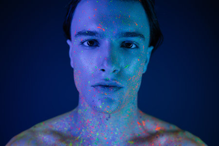 portrait of confident, self-assured man with bare shoulders posing in vibrant and colorful neon body paint while looking at camera on blue background with cyan lighting effectの写真素材