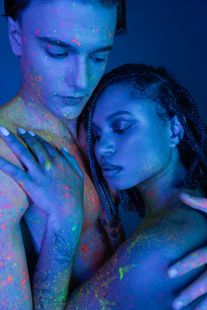 sensual interracial couple in colorful body paint embracing on blue background with cyan lighting, handsome bare-chested man and enchanting african american woman with dreadlocksの写真素材