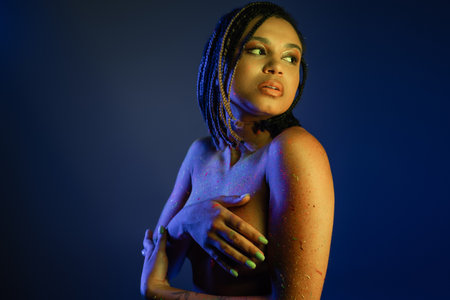 bare-chested african american woman with dreadlocks standing in colorful neon body paint, covering...