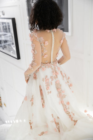 back view of brunette middle eastern woman with wavy hair walking in floral wedding dress inside of luxurious bridal salon, charming and elegant bride, blurred photography on white wallの写真素材