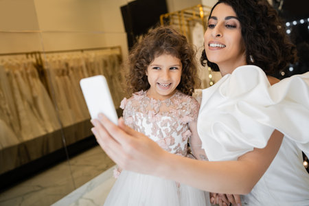 charming middle eastern bride with brunette hair in white wedding dress with puff sleeves and ruffles taking selfie on smartphone with happy daughter in bridal store with blurred backgroundの写真素材