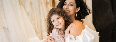 gorgeous middle eastern bride with brunette hair in white wedding dress with puff sleeves and ruffles embracing positive daughter in bridal store, white tulle fabrics on blurred background, bannerの写真素材
