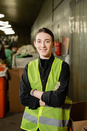 positive and young worker in gloves and high visibility jacket crossing arms and looking at camera...