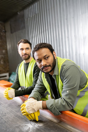 Indian sorter in protective vest and gloves looking away near conveyor and blurred colleague while...