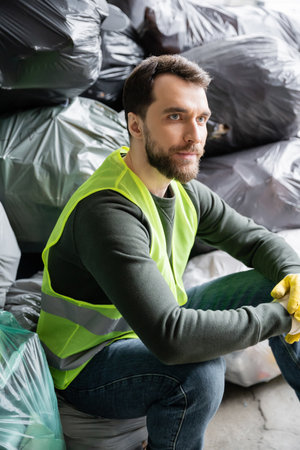 Bearded male worker in fluorescent vest and gloves looking away while sitting near blurred plastic bags with trash in garbage sorting center, recycling conceptの写真素材