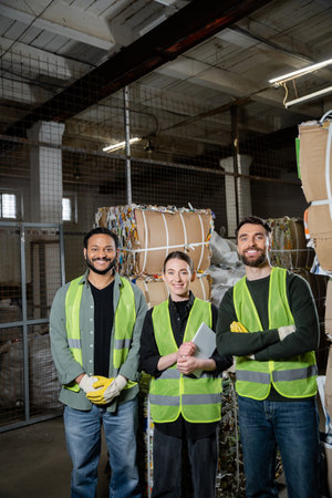 Smiling interracial sorters in protective vests holding digital tablet and looking at camera near...