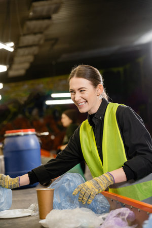 Cheerful young worker in protective vest and gloves sorting garbage on conveyor and looking away while working in waste disposal station, garbage sorting and recycling conceptの写真素材