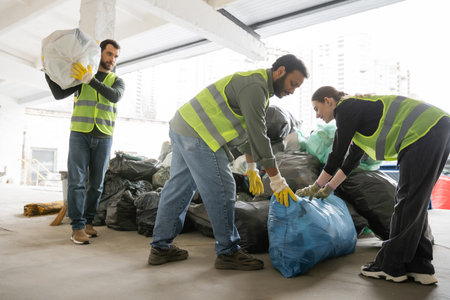 Smiling female worker in high visibility vest and gloves putting plastic bag with trash near...