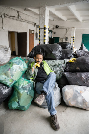 Confident indian worker in high visibility vest and gloves looking at camera while sitting on plastic bags with trash in garbage sorting center, recycling conceptの写真素材