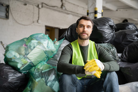 Bearded sorter in protective vest and gloves looking at camera while sitting near plastic bags with trash while working in blurred garbage sorting center, recycling conceptの写真素材