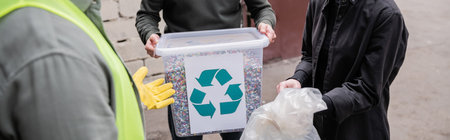 Cropped view of worker in protective vest and glove standing near volunteers with trash in outdoor waste disposal station, garbage sorting and recycling concept, bannerの写真素材