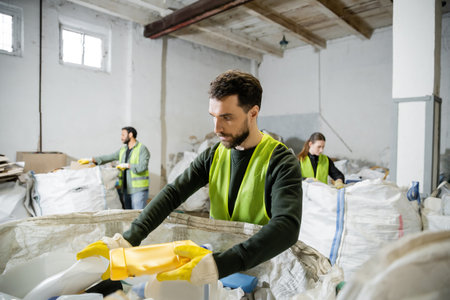 Bearded worker in protective gloves and vest taking plastic trash from sack while working near...