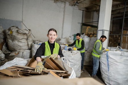 worker in gloves and high visibility vest looking at camera while putting cardboard in sack and working near multiethnic colleagues in waste disposal station, garbage sorting and recycling conceptの写真素材