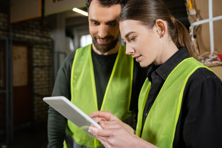 Young worker in safety reflective vest using digital tablet while standing near smiling colleague...