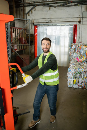 Smiling worker in safety vest and gloves pulling hand pallet truck while working near blurred waste...