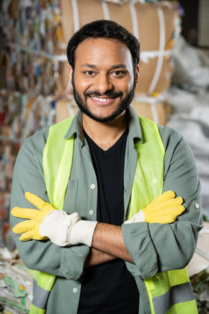 Cheerful indian worker in reflective vest and gloves looking at camera and crossing arms while standing near waste paper on blurred background in waste disposal station, garbage recycling conceptの写真素材