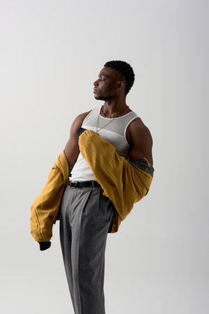Side view of good looking afroamerican model in bomber jacket and sleeveless t-shirt posing isolated on grey, contemporary shoot featuring stylish attire, muscularの写真素材