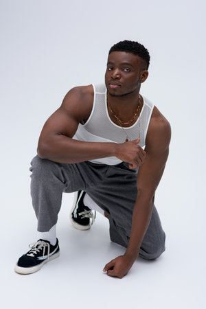 Full length of strong and young afroamerican model in tank top touching golden necklace and looking...