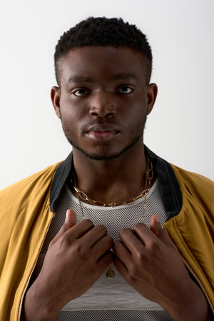Portrait of young and trendy afroamerican model in bomber jacket and golden necklaces looking at...