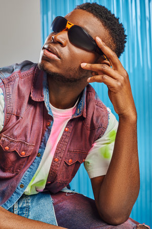 Portrait of stylish afroamerican model in sunglasses colorful denim vest and t-shirt posing on grey...