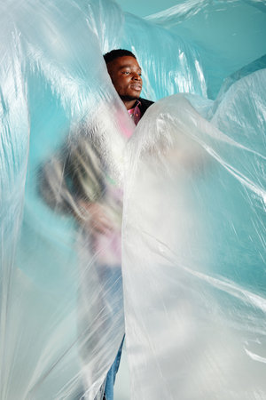 Young african american man with trendy hairstyle looking away while standing in cellophane on turquoise background, urban outfit and modern pose, creative expression, DIY clothingの写真素材