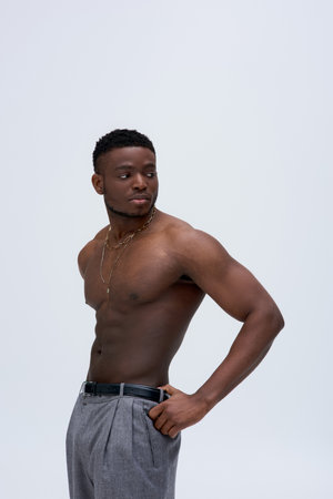 Muscular and sexy young afroamerican man in necklaces and pants standing isolated on grey, confident...