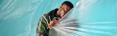Young and trendy afroamerican model in outwear jacket and led stripes covering face with cellophane on turquoise background, urban outfit and modern pose, banner, creative expression, DIY clothingの写真素材